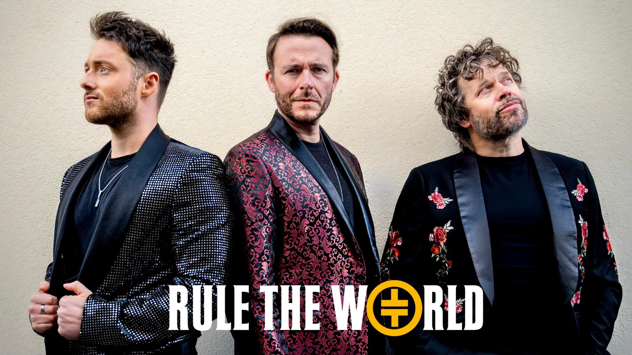 Three men - Rule the world standing next to eachother. The man in the middle is looking forward the camera while the other two look away.
